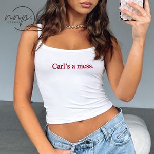 May include: White tank top with spaghetti straps. The text "Carl's a mess." is printed in red on the front. The top is paired with light blue denim jeans. A gold chain necklace is visible.
