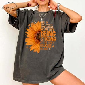 Może przedstawiać: Ciemnoszara koszulka z pomarańczową grafiką słonecznika i napisem "WE DON'T KNOW HOW STRONG WE ARE UNTIL BEING STRONG IS THE ONLY CHOICE WE HAVE." Koszulka ma wygląd vintage.