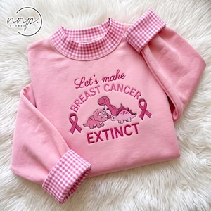 Op de afbeelding: Een lichtroze sweatshirt met een roze gingham kraag en mouwboorden. De voorzijde is voorzien van geborduurde tekst die luidt Let's make BREAST CANCER EXTINCT, vergezeld door drie roze dinosaurusillustraties en twee roze bewustzijnslinten.
