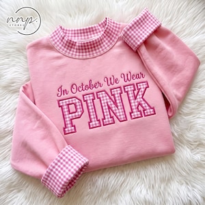 Op de afbeelding: Een lichtroze sweatshirt met een opstaande kraag en mouwboorden in een roze en wit vichy-ruitpatroon. De voorzijde toont de geborduurde tekst In October We Wear PINK, waarbij het woord PINK in een groot, blokvormig vichy-lettertype staat.