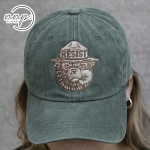 May include: A forest green baseball cap featuring an embroidered brown bear wearing a park ranger hat with the word RESIST stitched in white. The cap has a distressed, washed-out fabric texture and a curved brim with visible stitching details.