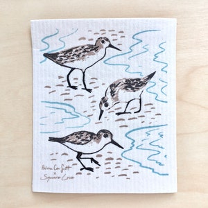 May include: A white cotton dishcloth with a blue and brown design of three sandpipers standing in water. The text "Beina Can Scott x Square Love" is printed in the bottom left corner.