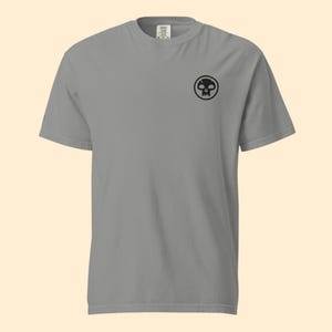 May include: A gray short-sleeved t-shirt featuring a black skull logo on the upper left chest. The shirt is made of a soft material and has a classic crew neck design. The shirt is a casual wear item.