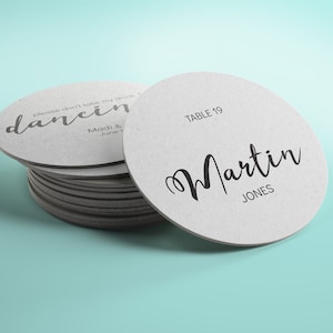 May include: A pile of white, circular coasters with grey edges. One coaster features the name "Martin JONES" and "TABLE 19" in black script. Another coaster reads "dancing" and "please don't take my drink" in a script font.