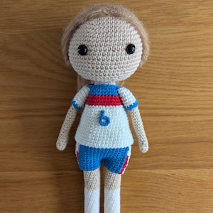 May include: Handmade crochet doll dressed in a white shirt with red and blue stripes, blue shorts, and green shoes. The doll has light brown hair and black eyes. The number 6 is on the shirt.