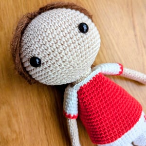 Könnte beinhalten: Handgemachte Häkelpuppe mit hellbeige Kopf, braunen Haaren und schwarzen Knopfaugen. Die Puppe trägt ein rot-weißes Outfit. Die Puppe liegt auf einer Holzoberfläche.