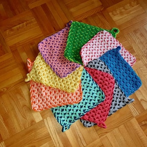 Könnte beinhalten: Ein Set aus neun gehäkelten Topflappen in verschiedenen Farben, darunter Grün, Rosa, Gelb, Orange, Blau und Grau. Die Topflappen sind in einem Kreis auf einem Holzboden angeordnet.