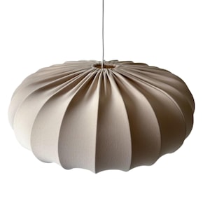 Op de afbeelding: Een beige hanglamp van stof. De lamp heeft een afgerond, geribbeld ontwerp, met een geplooid midden waar het snoer aan vastzit. Hij hangt aan een dunne, zilverkleurige draad tegen een witte achtergrond.