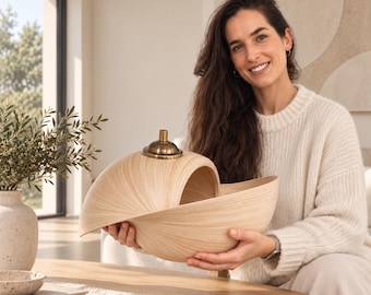 Op de afbeelding: Een lichtgekleurde, spiraalvormige houten kom met een glad, generfd oppervlak en een kleine, ronde, messingkleurige deksel of knop in het midden van de bovenste bocht. Het object heeft een gebogen, schelpachtige structuur met een open binnenkant.