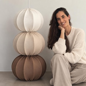 Peut inclure: Un lampadaire composé de trois abat-jours sphériques en tissu empilés, présentant un dégradé de couleurs allant du crème clair en haut au beige moyen au centre, et se terminant par un brun foncé à la base, suspendus par une tige verticale blanche.
