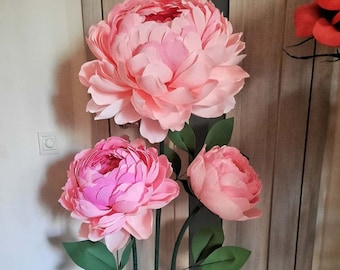 May include: Three large, handcrafted paper flowers in shades of light and dark pink, featuring layered, ruffled petals. The flowers are attached to tall, thin green stems with dark green leaves, all secured into a rectangular green base.