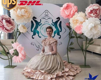 May include: A woman in a long, beige and pink dress with ruffles sits in front of a white backdrop with a blue and green logo. The backdrop is decorated with large, paper flowers in shades of pink and white. The logo features two stylized animals and the text "DHL". The text "Pray for Ukraine" is visible in the lower right corner of the image.