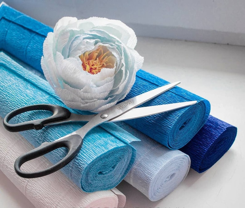 Pu&ograve; includere: Un fiore di carta bianco si trova sopra una pila di rotoli di carta crespa blu e bianca. Un paio di forbici con manici neri si trova sopra i rotoli.