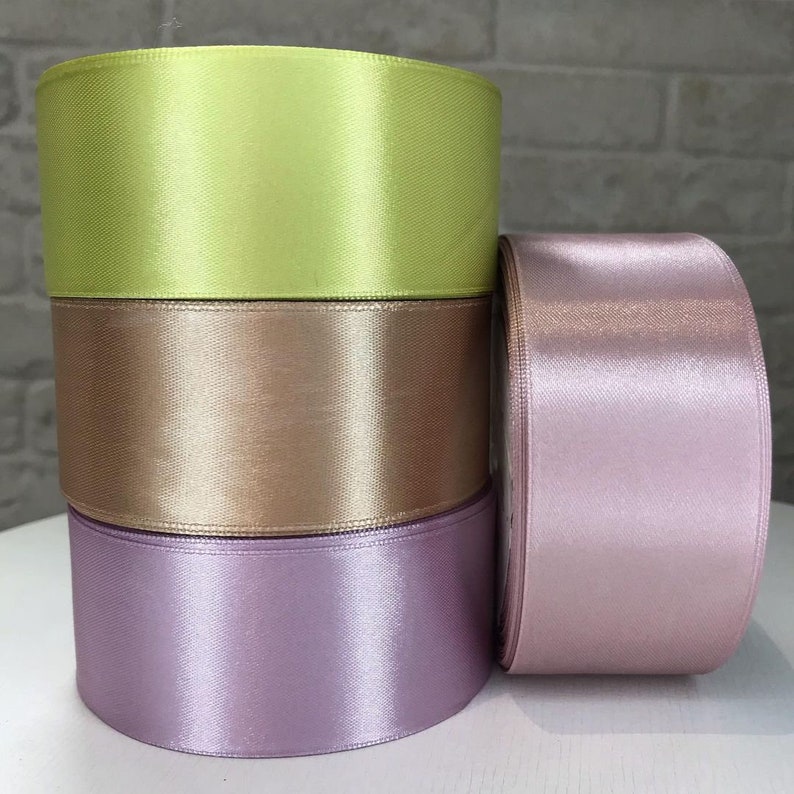 May include: Four spools of satin ribbon in different colors: light green, beige, light purple, and pink. The spools are stacked on top of each other.