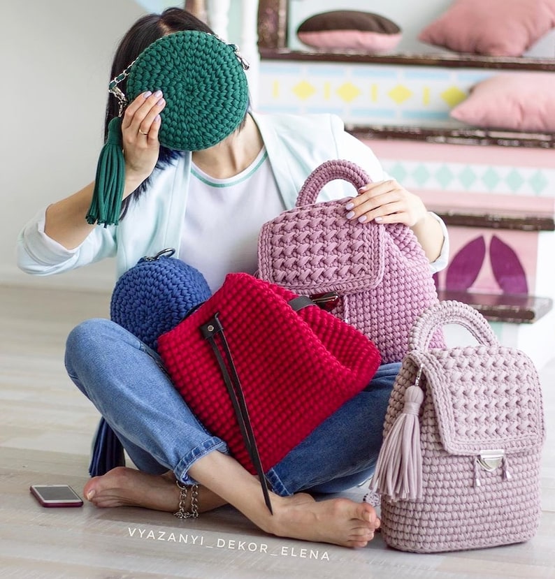 May include: A collection of four crocheted bags in different colors and styles. The bags include a round green bag, a blue bag, a red backpack, and a pink backpack. The bags are all made with a chunky yarn and have a textured look.