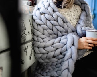 May include: A person is wrapped in a large, chunky knit gray blanket. The blanket is made of thick yarn and has a soft, cozy texture.