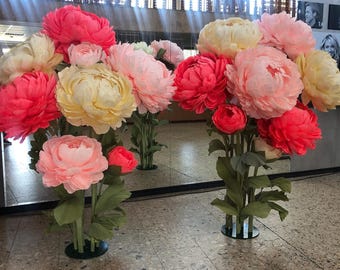 Puede incluir: Dos grandes arreglos florales con peonías de papel de gran tamaño en tonos coral vibrante, rosa suave y amarillo crema. Cada flor está unida a un tallo verde con hojas, apoyada sobre una base circular negra en el suelo.