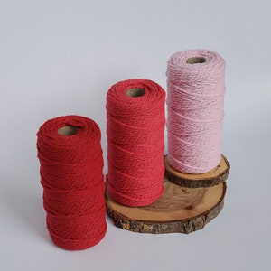May include: Three spools of macrame cord in shades of red and pink. The spools are displayed on small, round wooden slices against a white background. The cord is tightly wound, showing a textured surface.