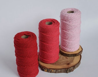 May include: Three spools of colorful macrame cord in shades of red and pink. The spools are displayed on small, round wooden slices against a white background. The cord is tightly wound, showing a textured surface.