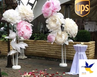 May include: White and pink paper flowers on white stands with red rose petals scattered on the ground. The background includes a brick wall and a sign with the UPS logo.