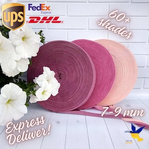 May include: Three spools of pink, red, and peach colored fabric ribbon. The ribbon is 7-9 mm wide and there are over 60 shades available. The ribbon is rolled up and the ends are visible. The ribbon is on a white surface with a white brick wall in the background.
