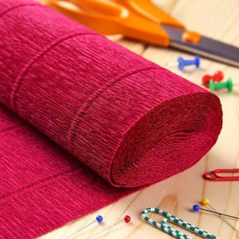 May include: A roll of deep magenta crepe paper with a distinct crinkled texture. The roll is positioned alongside orange-handled scissors, several colourful push pins in blue, red, and green, and a green and white patterned paper clip.