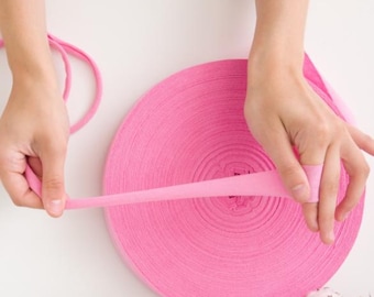 May include: A large roll of bright pink fabric ribbon, with a smaller length of the same material being held and stretched. The ribbon is tightly wound, creating a circular shape. The background is a plain white surface.