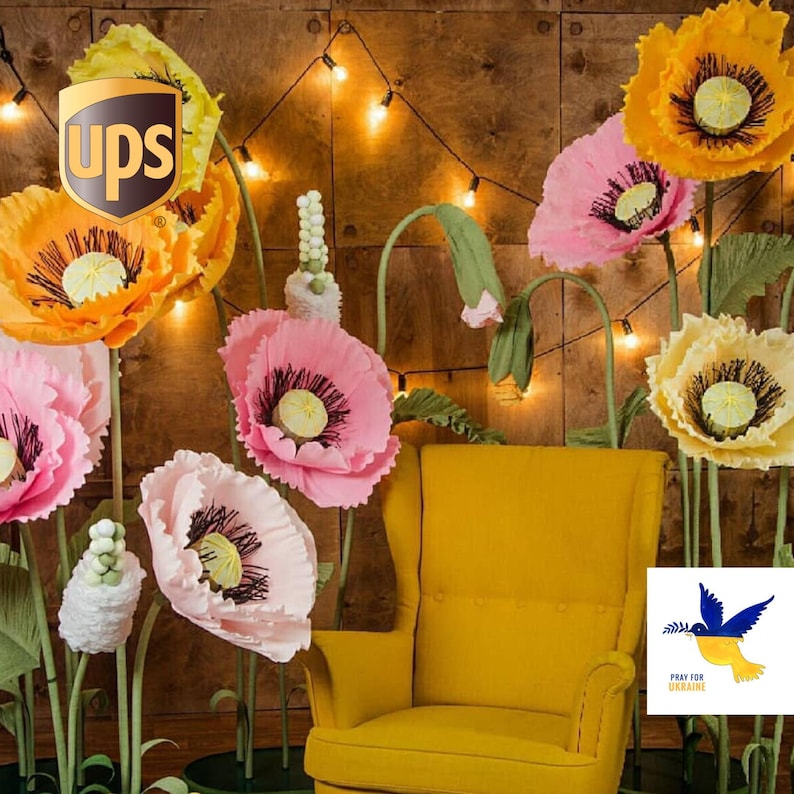 Giant handmade crepe paper poppy flowers in pink, yellow and white colors arranged around a yellow armchair with string lights, large wedding and event decoration.