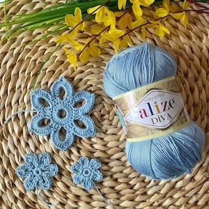 May include: A skein of light blue Alize Diva silky effect yarn next to several crocheted blue flowers. The yarn is on a woven, light brown surface, with yellow flowers and green stems in the background.
