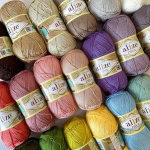 Op de afbeelding: Een verzameling kleurrijke Alize Diva garenbollen. Het garen is verkrijgbaar in verschillende kleuren, waaronder bruin, beige, roze, paars, geel, groen en blauw. Elke bol is verpakt met een label met de merknaam en productinformatie.