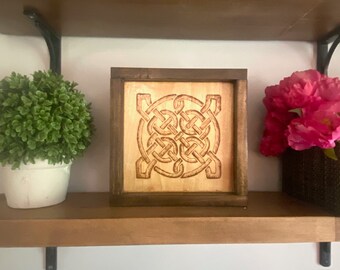 May include: A wooden framed art piece with a Celtic knot design. The artwork is a square shape with a light wood center and a dark brown frame. The piece is displayed on a wooden shelf with a small green plant and pink flowers.