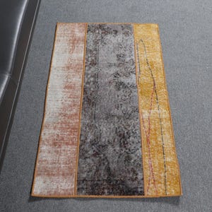 Op de afbeelding: Een rechthoekig tapijt met drie verticale panelen. Het linker paneel is lichtbeige met bruine en witte patronen. Het middelste paneel is donkergrijs met een vervaagd bloemenpatroon. Het rechter paneel is geel met rode en zwarte lijnen.