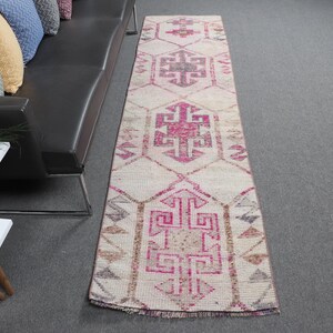 Puede incluir: Una alfombra rectangular de estilo vintage con fondo crema y patrones geométricos en tonos rosa, marrón y gris. La alfombra está sobre una superficie gris y presenta un diseño central con motivos repetidos.