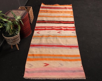 May include: A long, rectangular woven runner rug featuring horizontal stripes in shades of orange, red, pink, brown, and cream. The rug includes small, repeating geometric arrow-like motifs scattered across the surface and has a fringed edge.