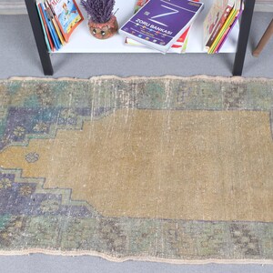 May include: A rectangular area rug featuring a central tan mihrab design on a field of muted green, blue, and brown tones. The rug has a stepped arch pattern, a distressed texture, and white fringe along the short edges, showcasing a worn appearance.