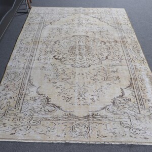 May include: A large, rectangular beige rug with an intricate, faded brown floral pattern. The rug has a central medallion design and a detailed border. The rug is placed on a gray surface.