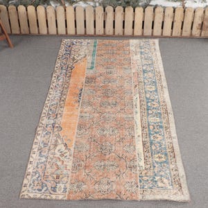 Puede incluir: Una alfombra rectangular de estilo vintage con un aspecto desgastado. La alfombra presenta un campo central de tonos cálidos naranja y beige, bordeado por intrincados patrones azules y crema. La alfombra está colocada sobre una superficie gris.