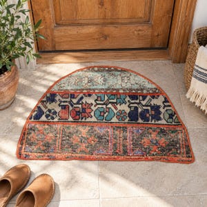 Può includere: Uno zerbino semicircolare caratterizzato da un design patchwork a tre fasce orizzontali distinte. La fascia superiore è azzurra con motivi floreali, quella centrale mostra fiori blu e rossi, mentre quella inferiore ha un motivo geometrico scuro.
