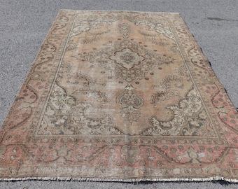 May include: A rectangular area rug featuring a central medallion design in shades of tan, beige, and muted brown. The border displays a repeating floral pattern in soft pink and grey tones, with a fringed edge along the bottom of the rug.