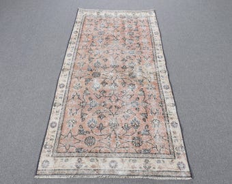 May include: A rectangular area rug featuring a faded peach-colored center field with a dark gray and cream floral vine pattern. The rug has a wide cream-colored border with repeating circular floral motifs and short fringe on both ends.