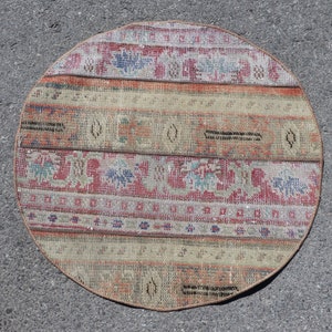 Puede incluir: Una alfombra circular con un diseño de patchwork que presenta bandas horizontales de diversos patrones. La paleta de colores incluye tonos apagados de rojo, beige, azul y gris. La alfombra tiene un borde acabado y un aspecto desgastado.