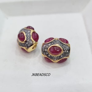 May include: Two gold-toned beads with a red gemstone and diamond accents. The beads are oval-shaped and have a textured surface.  The beads are set against a white background.