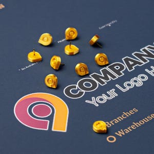 May include: A collection of small, gold-colored push pins scattered across a dark blue surface featuring a printed graphic design. The design includes the text COMPANY, Your Logo Here, Branches, and Warehouse, alongside a stylized letter a logo.