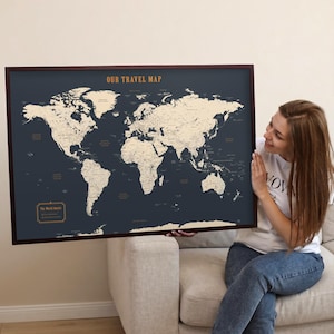May include: A framed world map with a navy blue background and a dark brown frame. The map shows a cream-coloured world map with country outlines. The text "OUR TRAVEL MAP" is at the top. The map is being held by a person.