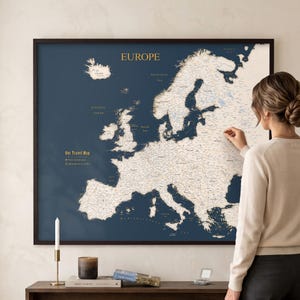 Peut inclure: Une carte murale encadrée de l'Europe présentant un fond bleu foncé avec des terres de couleur crème. La carte affiche le texte "EUROPE" en haut et "Our Travel Map" avec une légende sur le côté gauche. La carte est dans un cadre sombre.