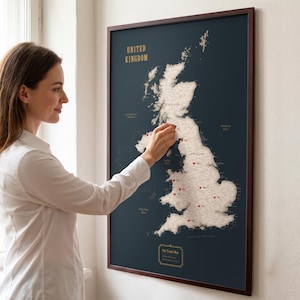 Könnte beinhalten: Eine gerahmte Wandkarte des Vereinigten Königreichs mit dunkelblauem Hintergrund und einer hellen Karte der Britischen Inseln. Die Karte enthält den goldenen Schriftzug UNITED KINGDOM und mehrere rote Stecknadeln an verschiedenen Orten.