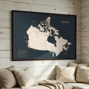 May include: A framed wall map of Canada featuring a dark blue background with a light beige landmass. The map displays geographical details, including bodies of water and regional labels, with the word CANADA printed in gold in the top right corner.