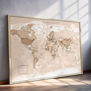 May include: A framed world map with a vintage aesthetic. The map features a beige background with brown landmasses and is labeled "Our Adventure Map." The frame is a light beige color. The map includes the text "The Oakland Family" and a list of places.