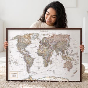 May include: A framed world map titled TRAVEL MAP with a muted color palette of beige, brown, and soft blue tones. The map features a legend box in the bottom left corner labeled Our Adventures with two bullet points reading Rachel's trips and Viley's trips.
