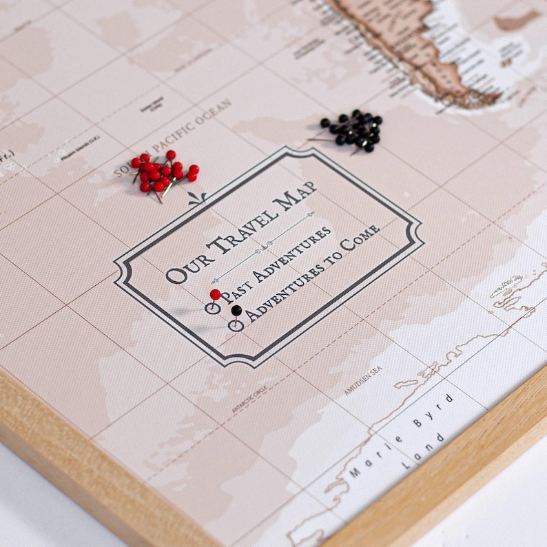 May include: A framed world map with the text "OUR TRAVEL MAP" and the phrases "PAST ADVENTURES" and "ADVENTURES TO COME." Red and black push pins mark locations. The map is in neutral tones with a wooden frame.